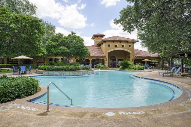 Apartments in Stone Oak San Antonio TX Outdoor swimming pool surrounded by greenery and lounge chairs, with a large building featuring a red-tiled roof in the background under a partly cloudy sky. The Montecristo Apartments in San Antonio North Loop San Antonio, TX 78232 p: (210) 672-4919 f: (210) 877-5455